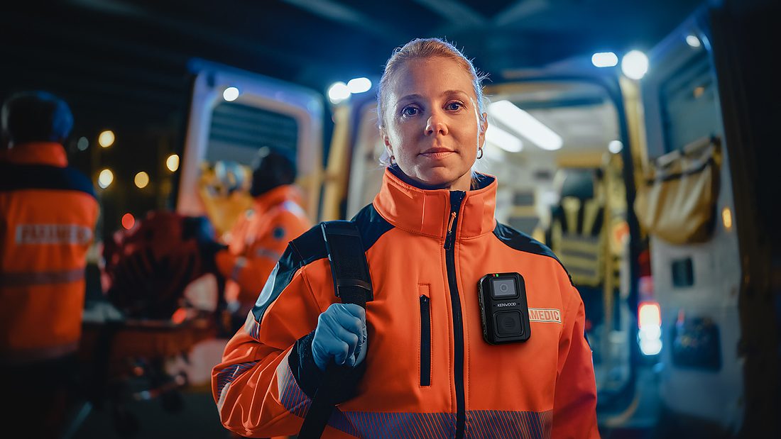 Paramedic wearing a KENWOOD body camera on her jacket beside an ambulance